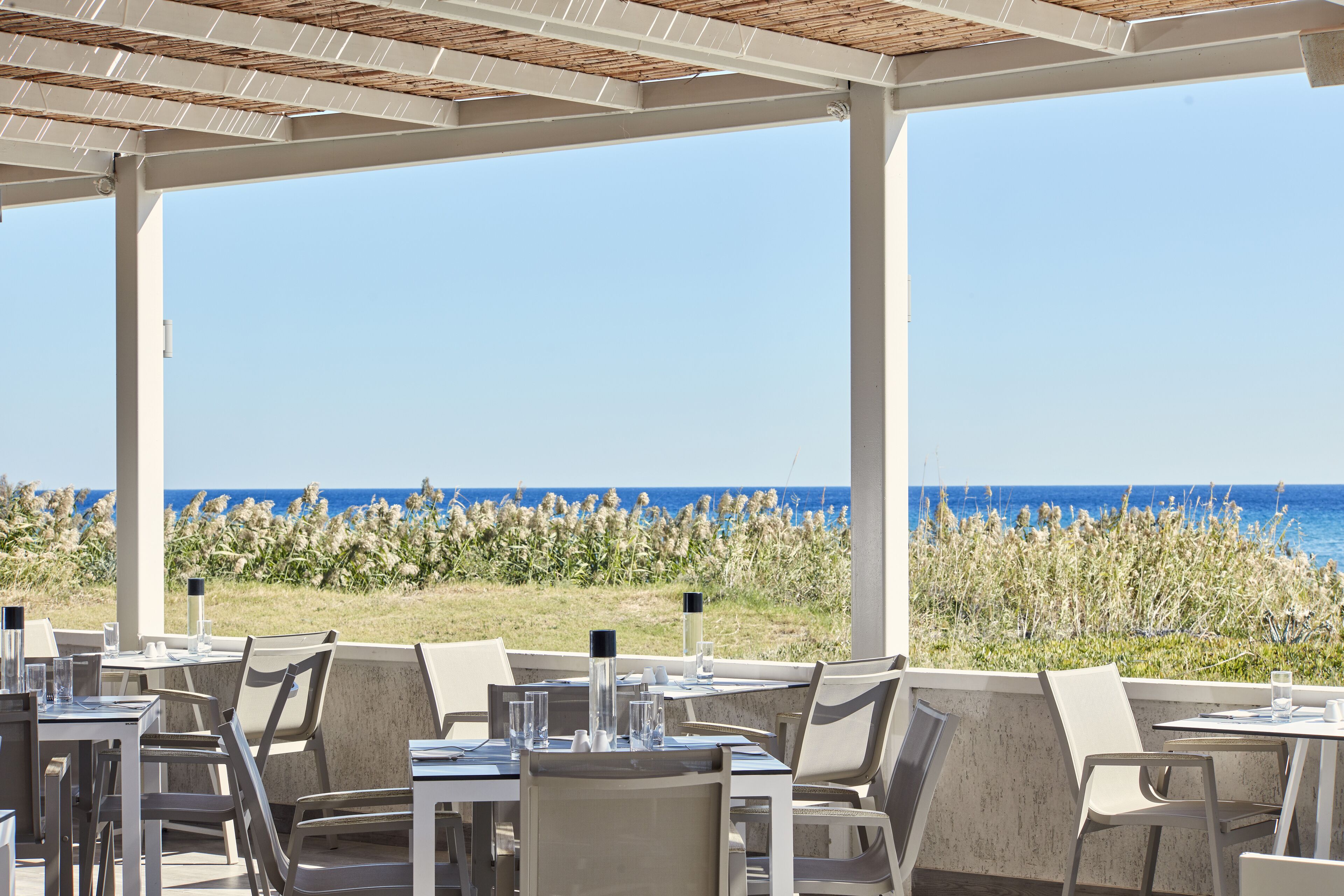 A seaside restaurant with tables and chairs, a pergola, and a view of the ocean.