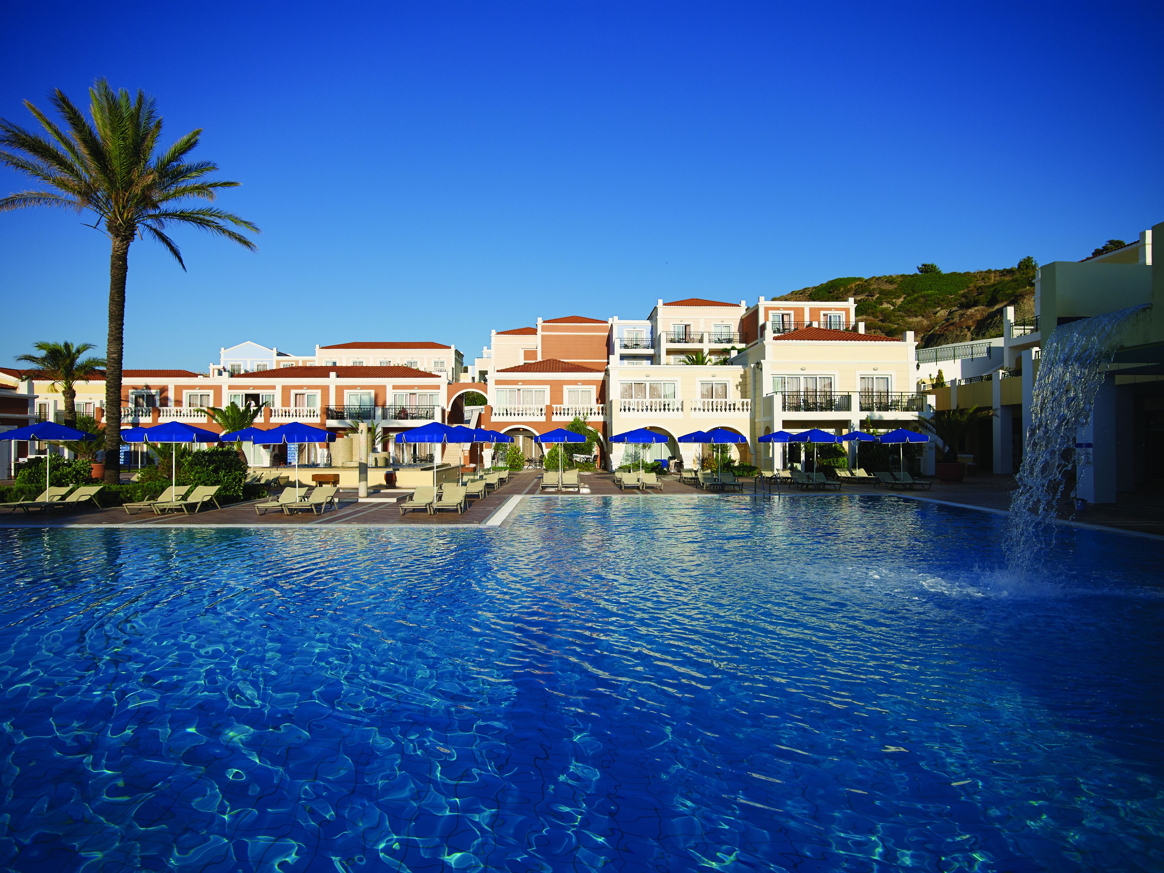 Ein Hotelpool mit klarem, blauem Wasser, Liegestühlen und blauen Sonnenschirmen.