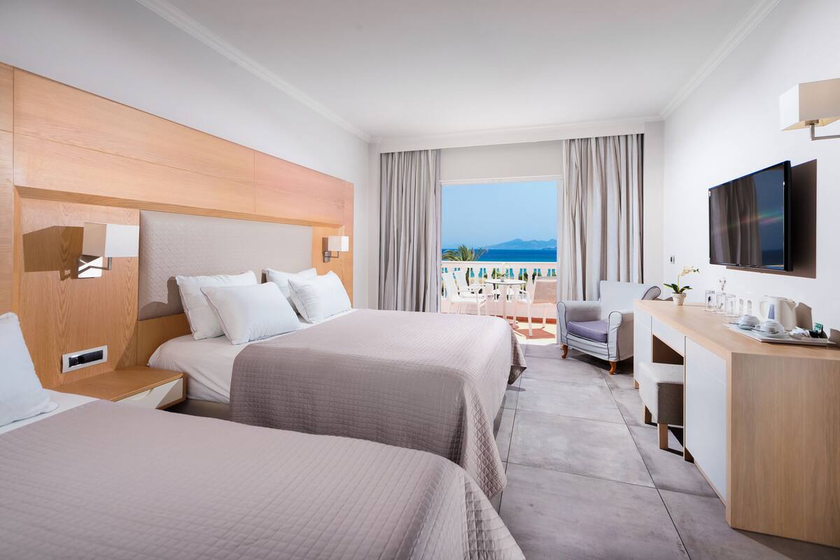 A hotel room with two beds, a desk, a TV, and a balcony with a view of the sea.