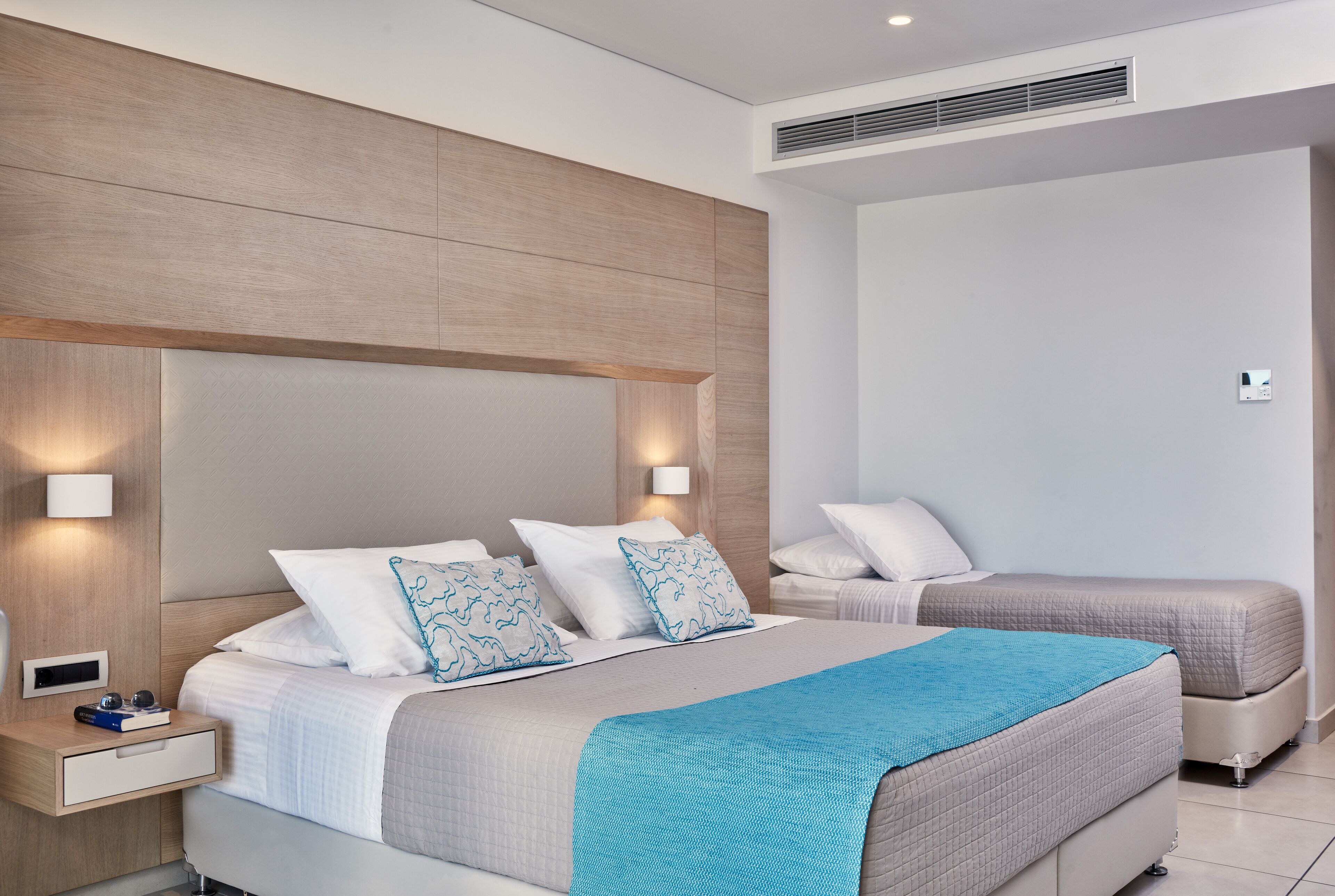 A modern hotel room with a large bed, a smaller bed, a nightstand with a drawer, and a wall-mounted light.