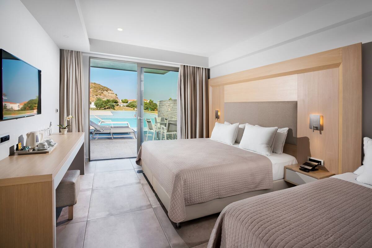 A hotel room with two beds, a desk, a TV, and a view of a pool and mountains.