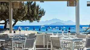 Area pranzo all'aperto con tavoli e sedie bianche, affacciata su una spiaggia con ombrelloni e montagne sullo sfondo.