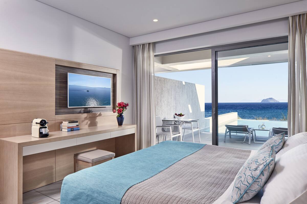 A modern hotel room with a large bed, a desk with a flat-screen TV, a small table with chairs, and a balcony with a view of the sea.