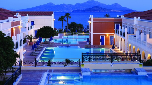 Un resort con una piscina centrale, numerosi balconi e vista sul mare e sulle montagne.