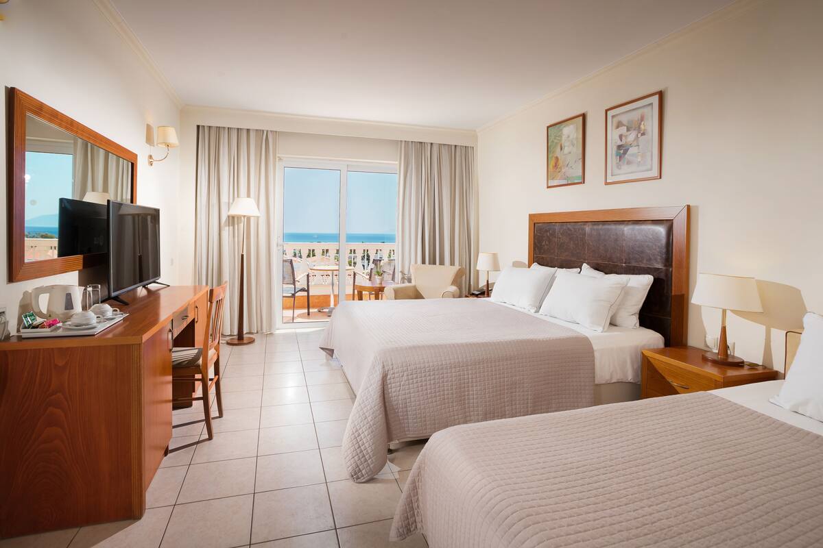 A hotel room with two beds, a desk with a TV, and a balcony with a view.