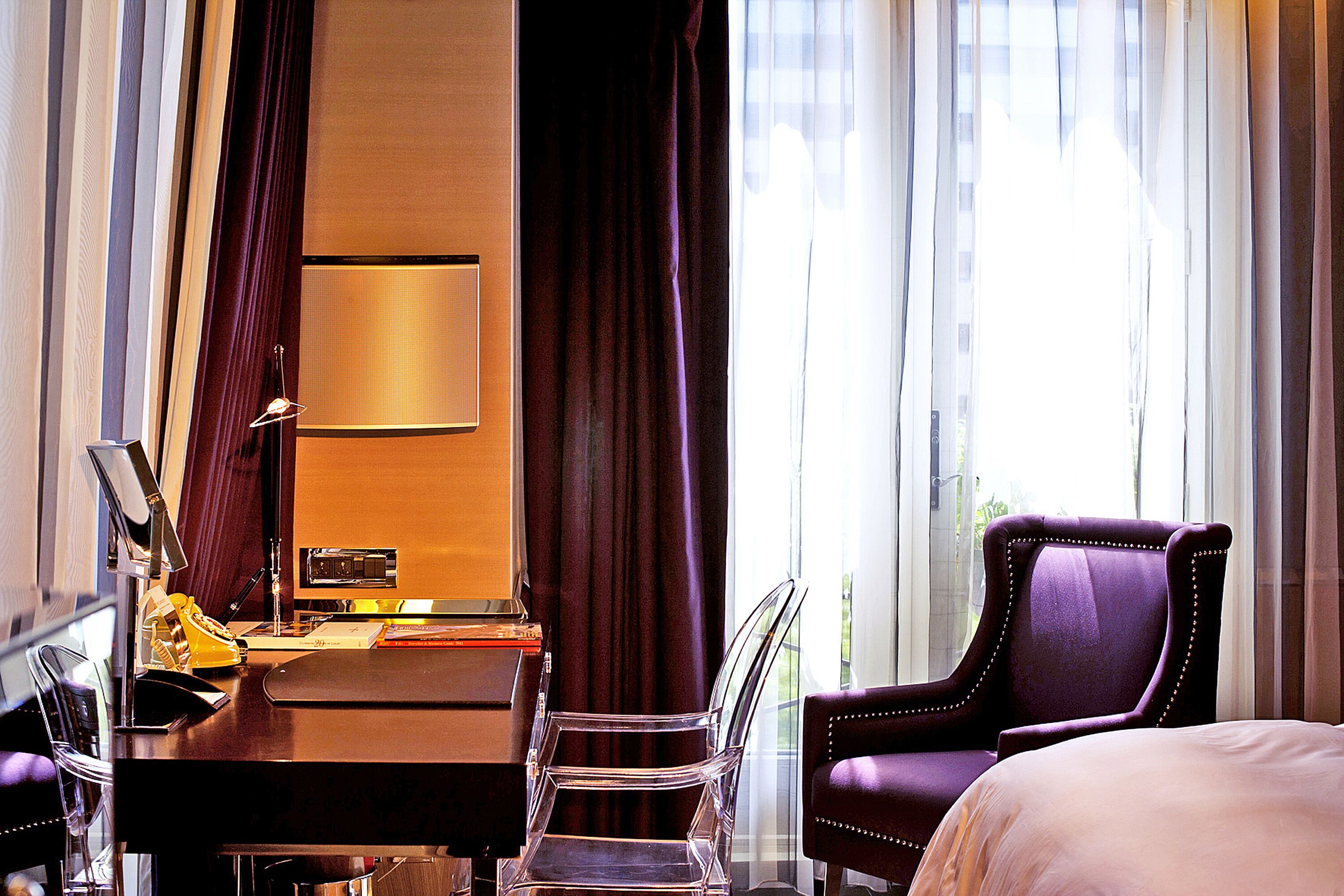 A hotel room with a bed, a desk with a lamp, a chair, and a large window with curtains.