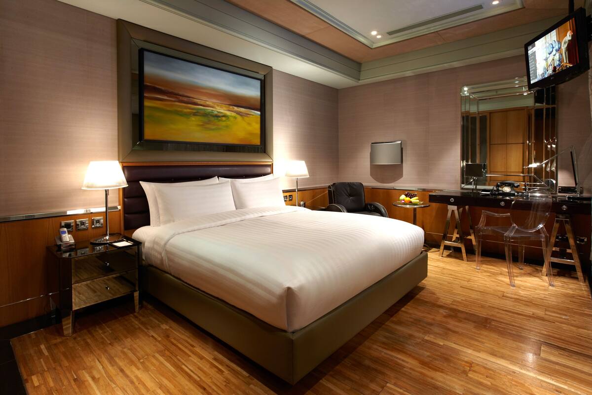 A hotel room with a large bed, bedside lamps, a desk with a chair, a television, and a painting on the wall.