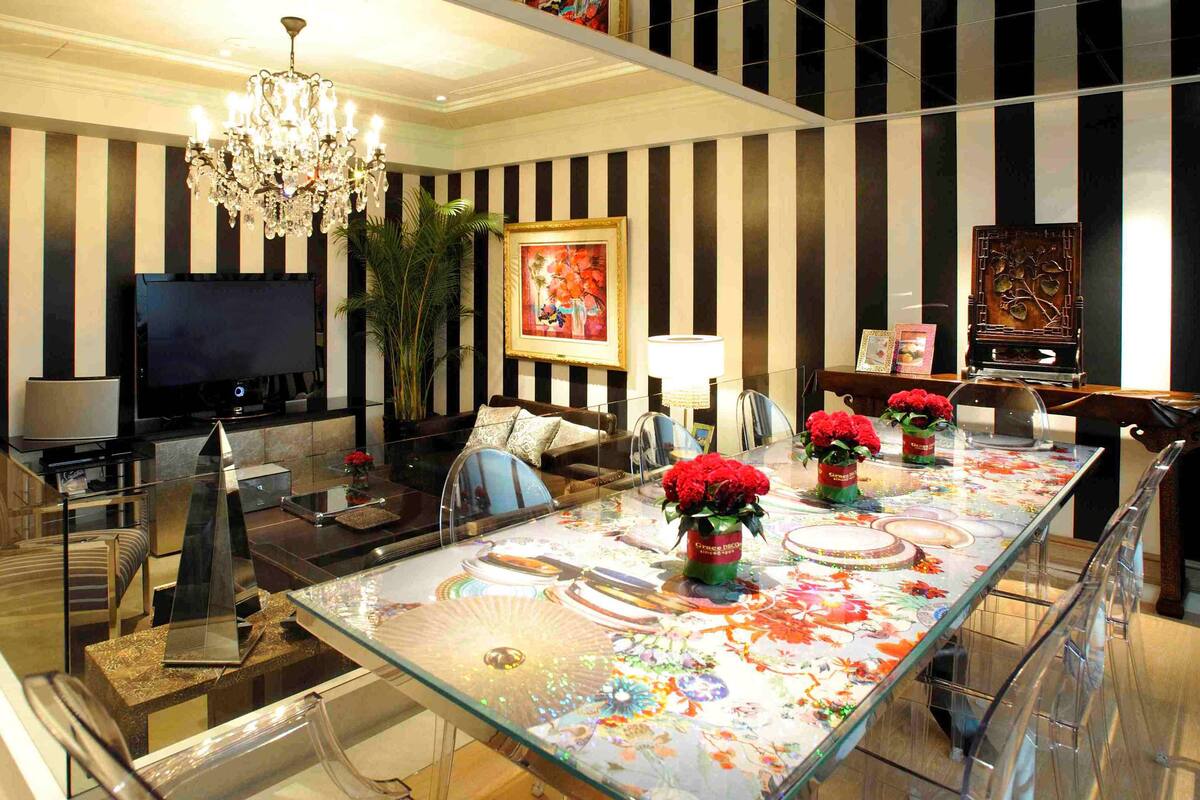 A modern living room with a large TV, comfortable seating, a chandelier, and a dining area with a glass table and red flowers.