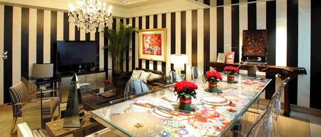 A modern living room with a large TV, comfortable seating, a chandelier, and a dining area with a glass table and red flowers.