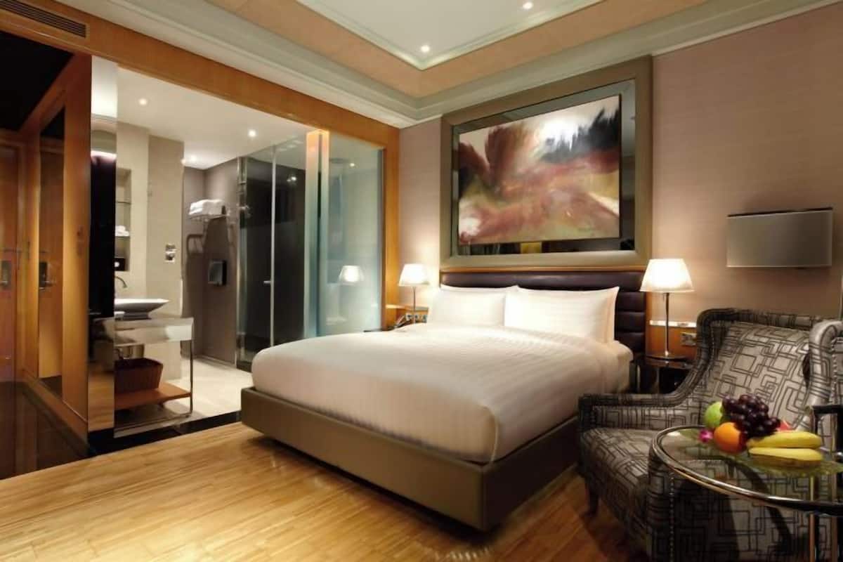 A hotel room with a large bed, a sitting area with a chair and a small table, a large painting on the wall, and a bathroom visible through an open door.