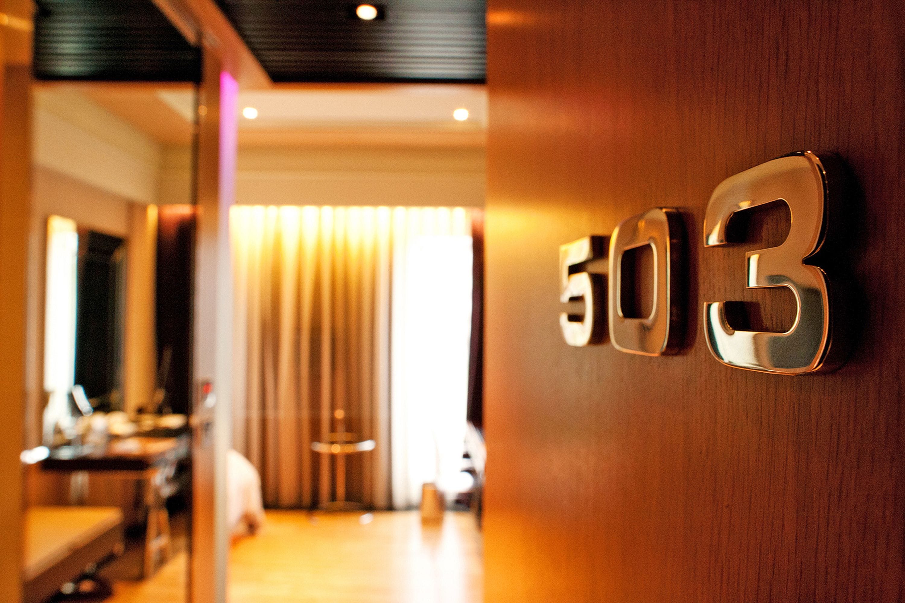 Interior de un hotel con un cartel con el número '503' en la pared, suelo de madera y una persona caminando al fondo.