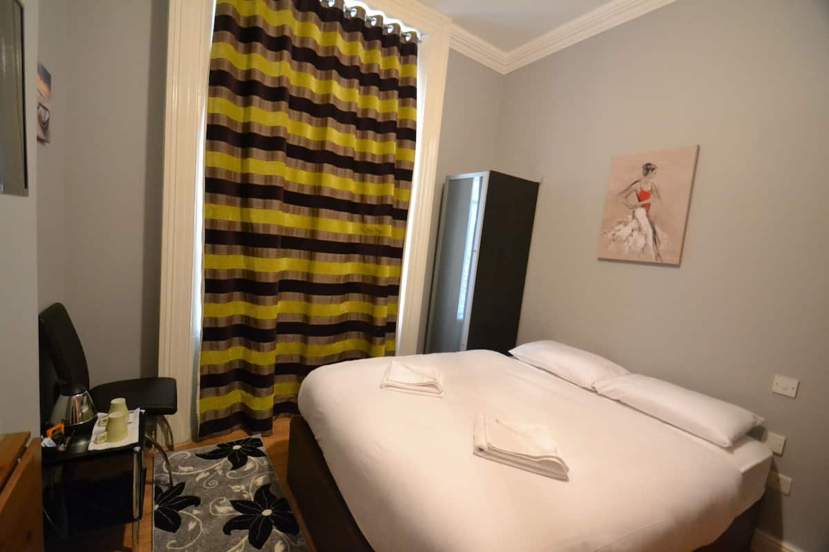 Ein Hotelzimmer mit Bett, Stuhl, Schreibtisch mit Lampe, einem gestreiften Vorhang und einem Bild an der Wand.