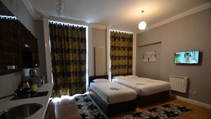 A modern hotel room with a kitchenette, two beds, a TV, and a large window with curtains.