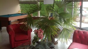A room with a pool table, red upholstered chairs, a large potted plant, and a window with a view of greenery. - Brit Hotel le Magdalena (Varennes-Vauzelles)