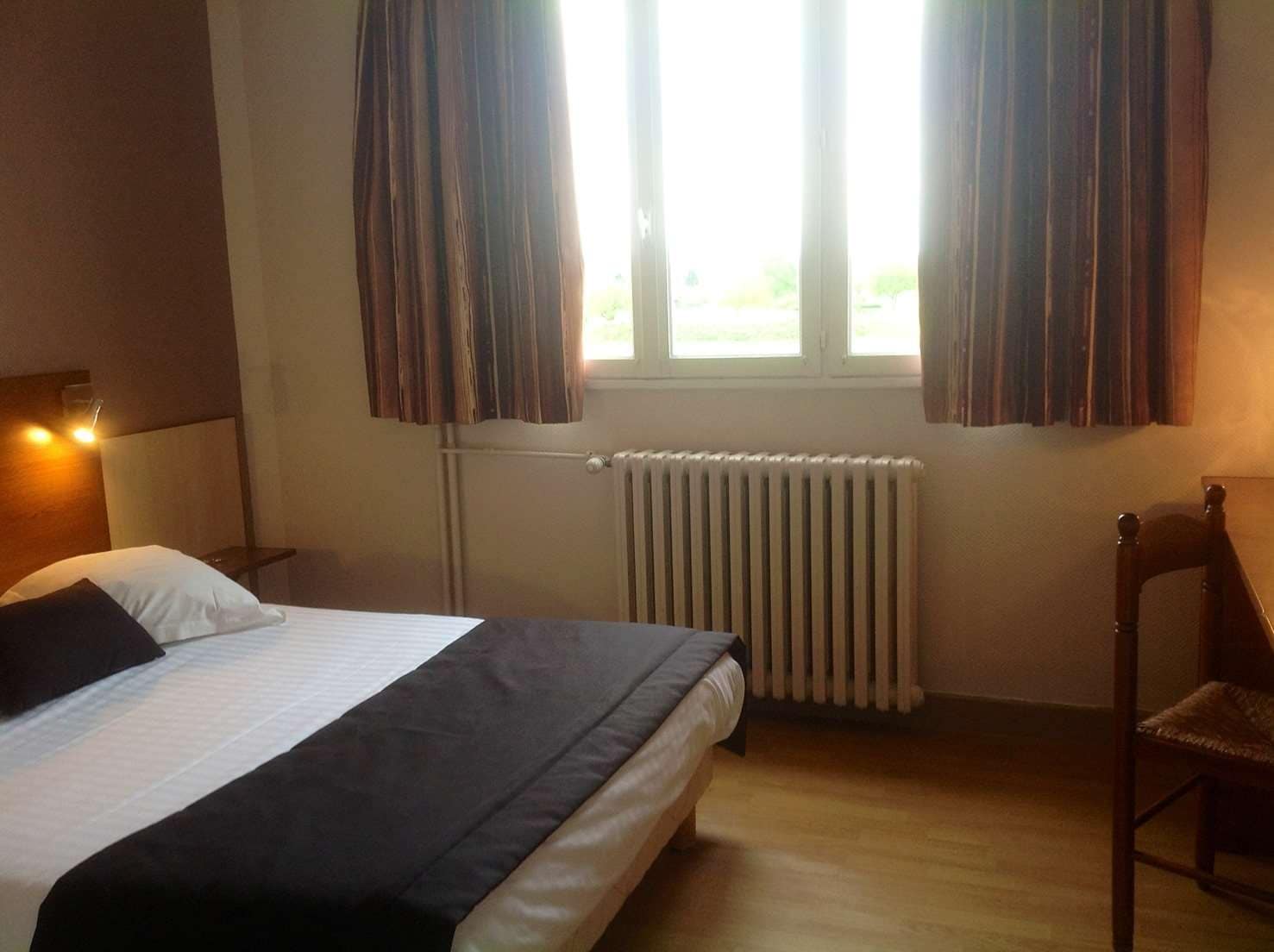 Ein Hotelzimmer mit einem Bett, einem Sessel, einem Nachttisch, einem Fenster mit Vorhängen und einer Heizung.