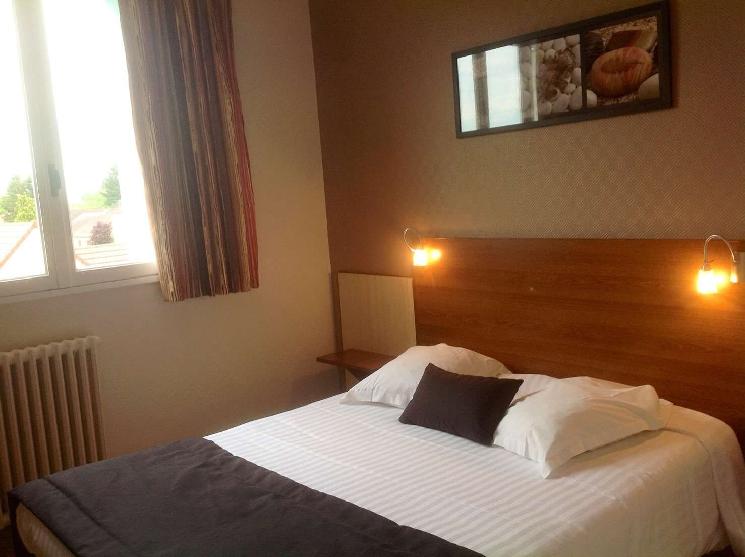 Habitació d'hotel amb un llit, una tauleta de nit, una làmpada, una finestra amb cortines i un quadre penjat a la paret.