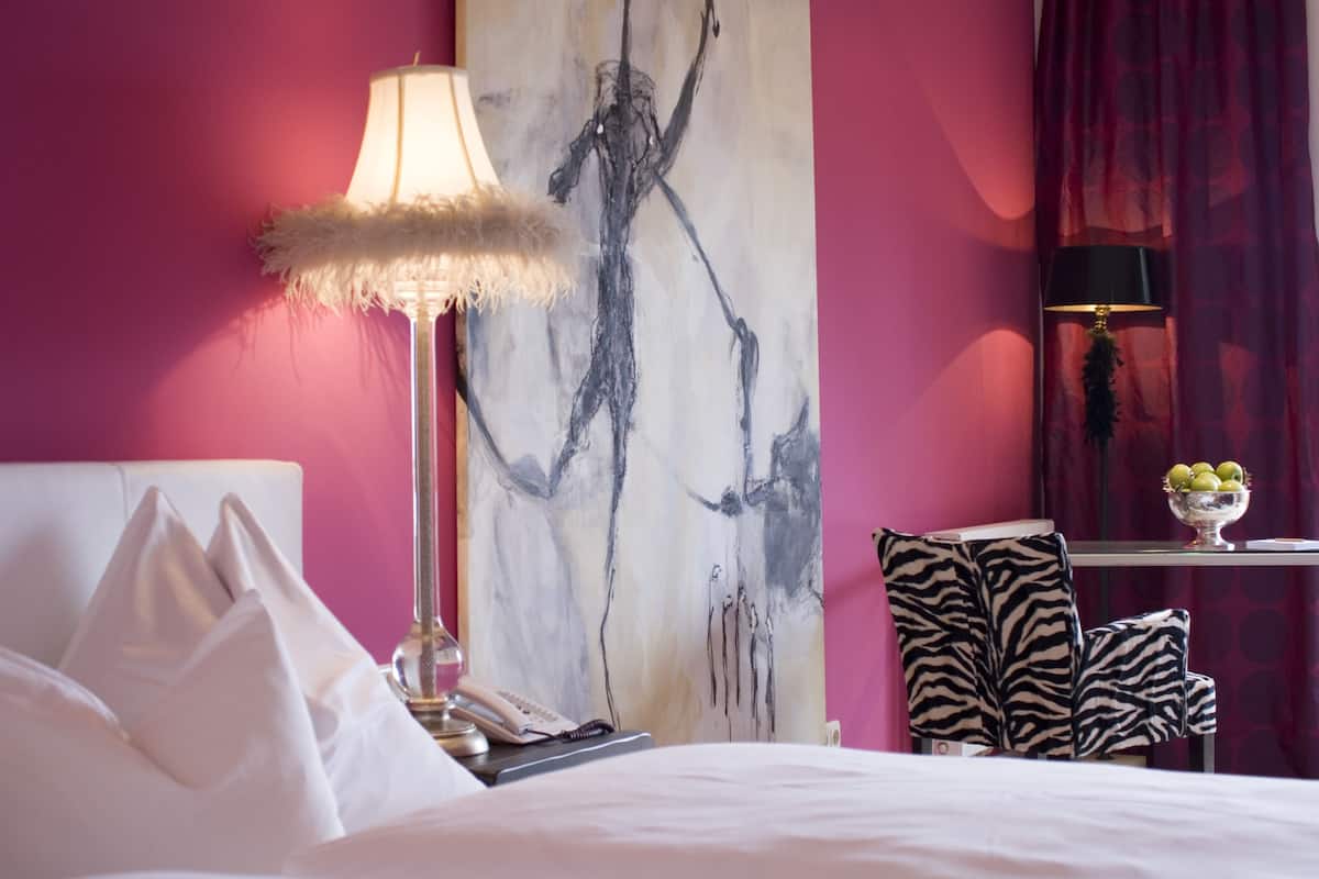 A hotel room with a bed, a large abstract painting, a zebra-patterned chair, a table with a bowl of fruit, and a lamp with a feathered shade.
