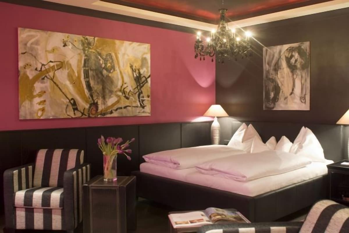A hotel room with a bed, a chair, a nightstand, and two paintings on the wall.