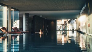 An indoor swimming pool with clear water, sun loungers, and a view of buildings through large windows.