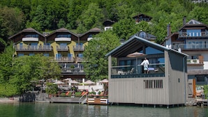 A wooden floating structure with a balcony, surrounded by a lake and multi-story buildings in the background.