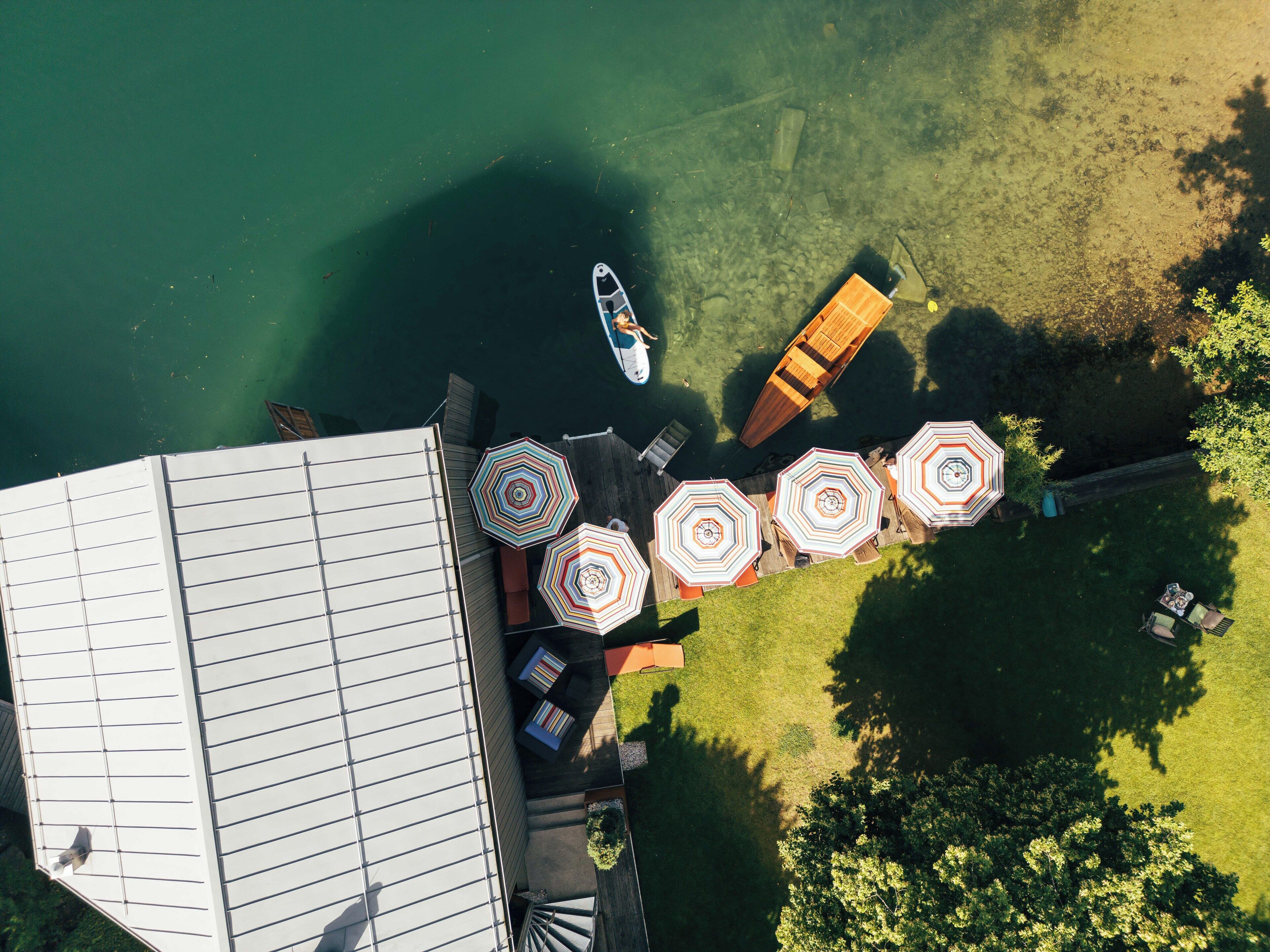 An aerial view of a lakeside property with a dock, a boat, and outdoor seating with umbrellas.