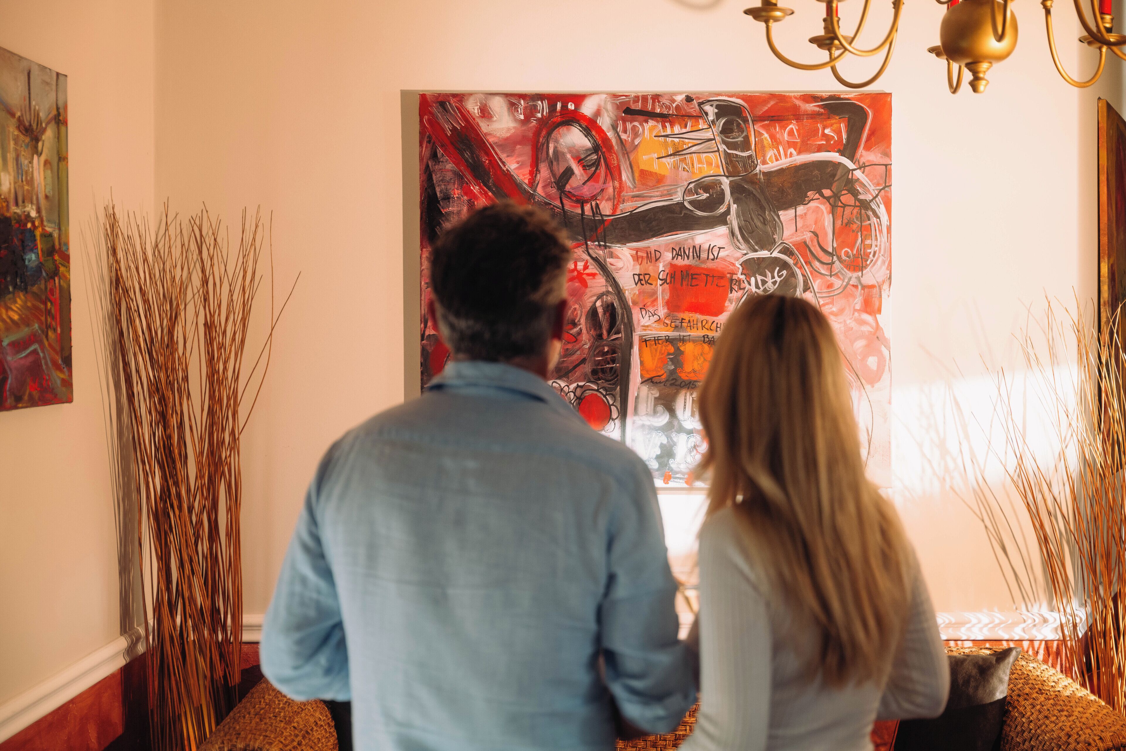 A room with a large abstract painting on the wall, a chandelier, and tall decorative plants.