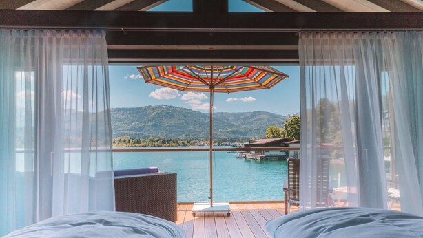 A bedroom with a large window overlooking a lake, a colorful striped umbrella, and a wooden deck with outdoor furniture.