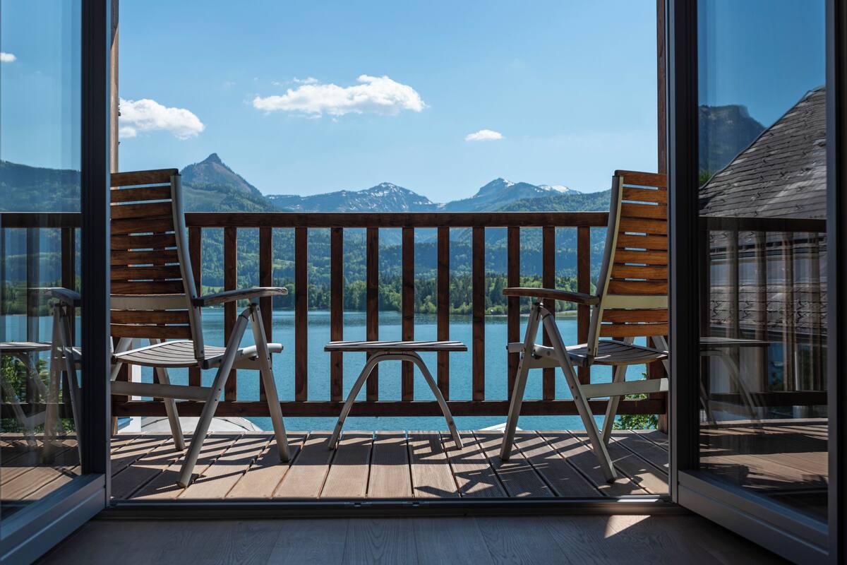 A balcony with wooden chairs and a table, overlooking a lake and mountains.