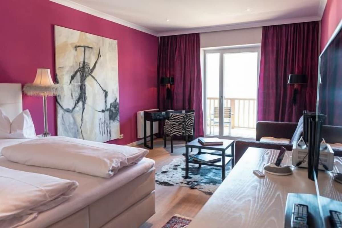A hotel room with a bed, desk, chair, artwork, and a view of the outside.