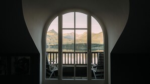 A room with a large arched window overlooking a lake and mountains. There are two chairs outside the window.