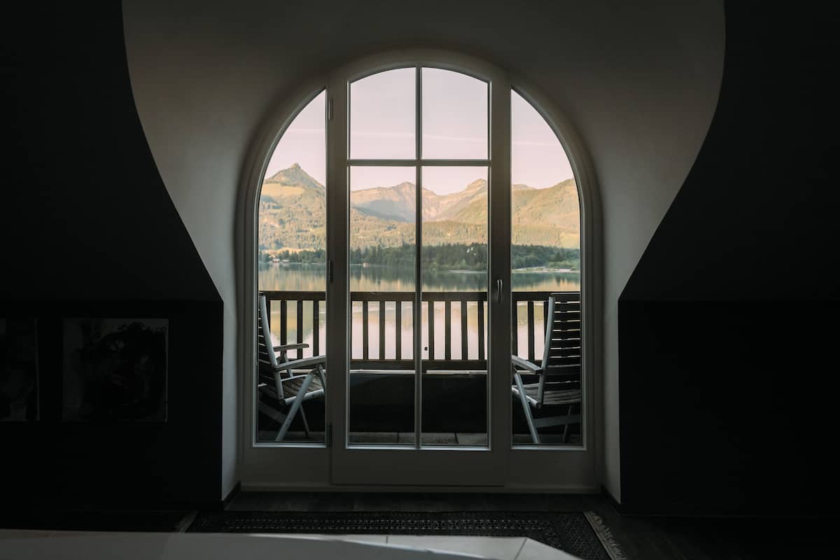 A room with a large arched window overlooking a lake and mountains. There are two chairs outside the window.