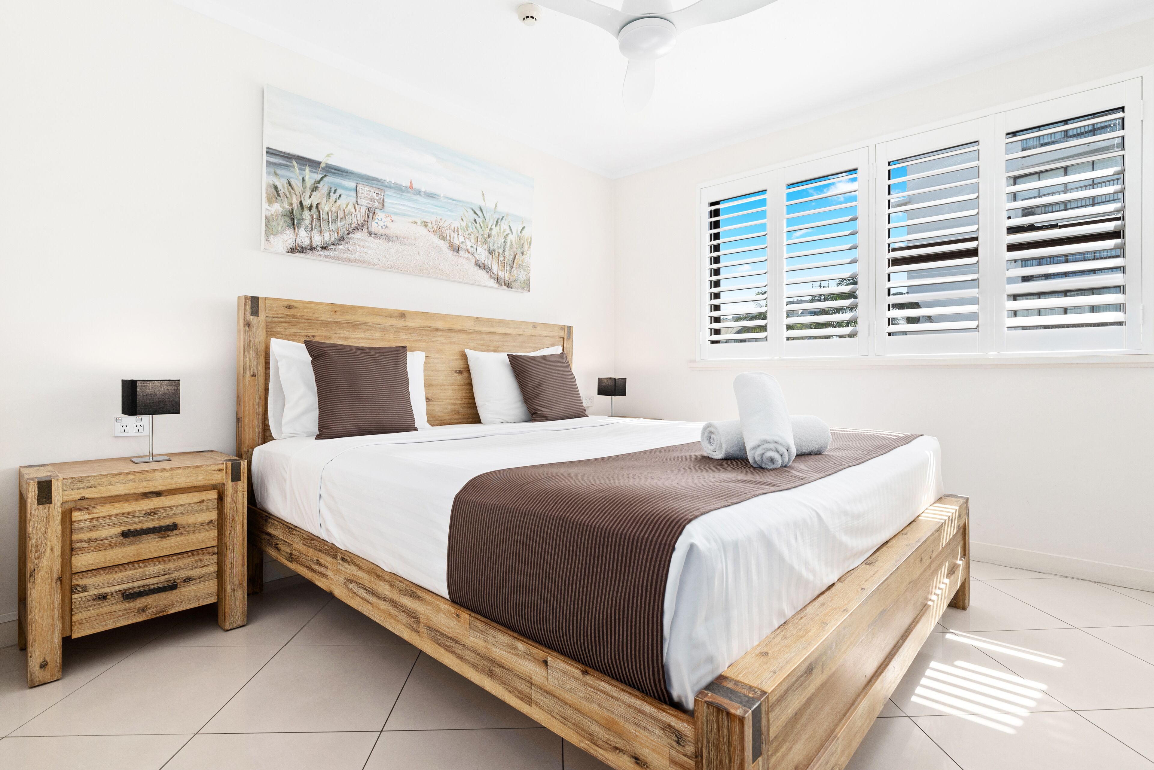 Een slaapkamer met een houten bed, witte beddengoed, een nachtkastje, een schilderij aan de muur en een raam met jaloezieën.