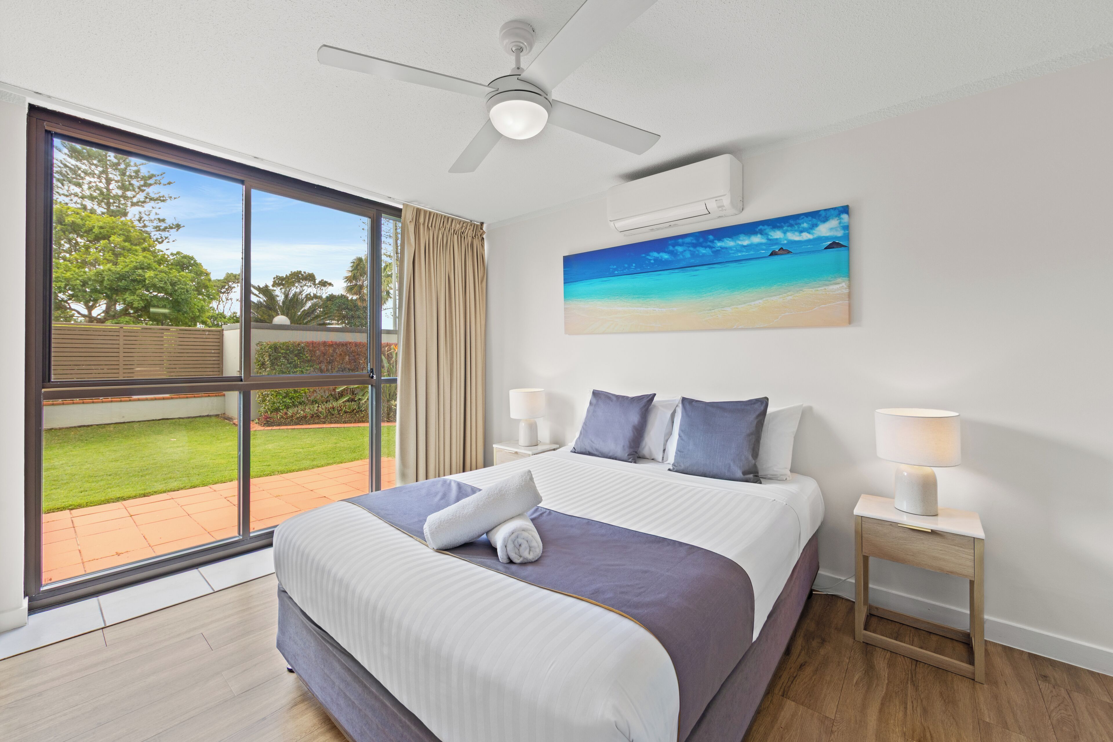 A modern hotel room with a large bed, a ceiling fan, a painting of a beach, and a view of a garden through a sliding door.