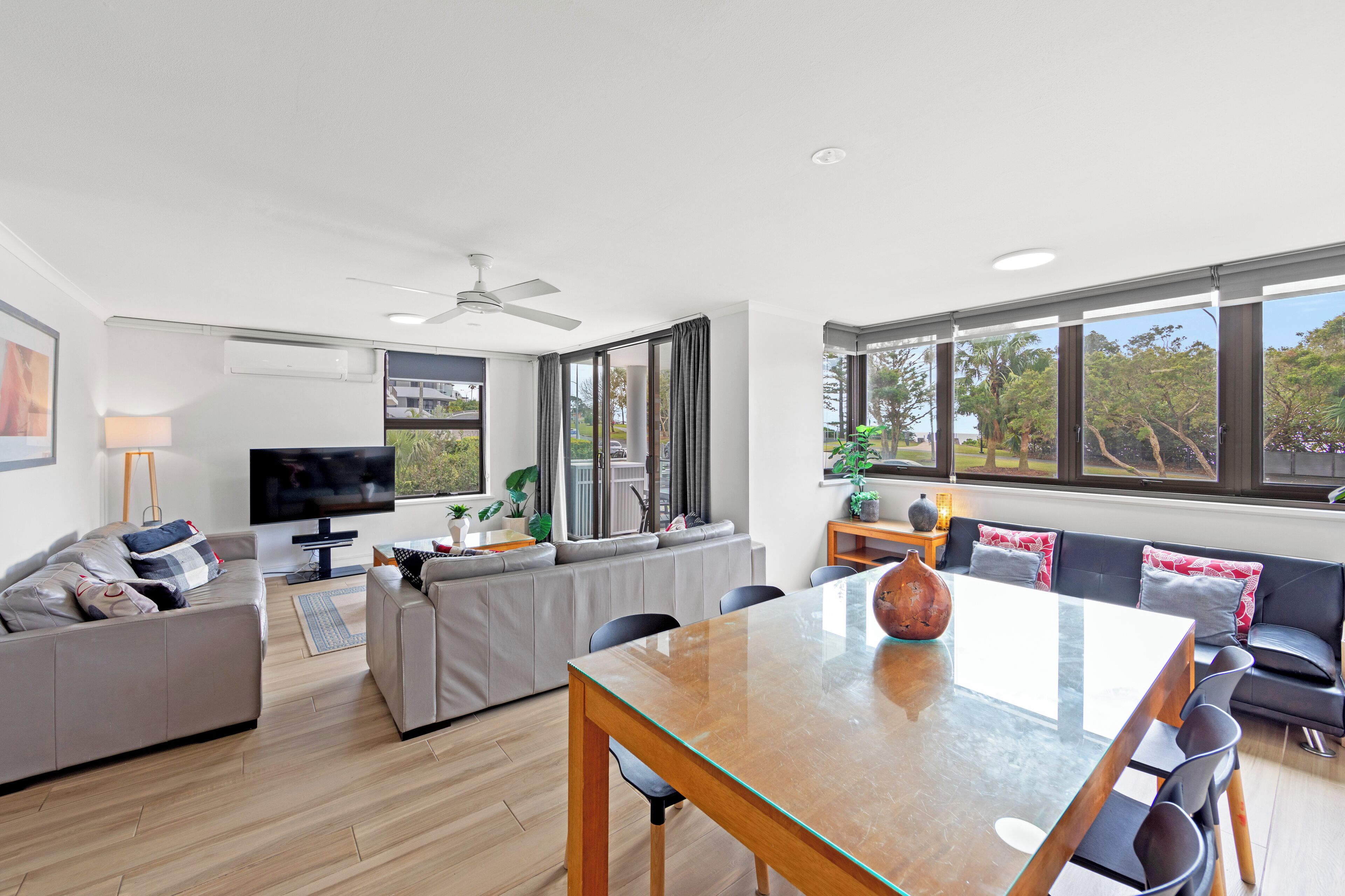 A modern living room with a glass dining table, grey sofas, a flat-screen TV, and large windows offering a view of greenery.