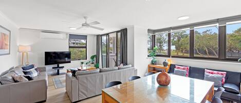 A modern living room with a glass dining table, grey sofas, a flat-screen TV, and large windows offering a view of greenery.