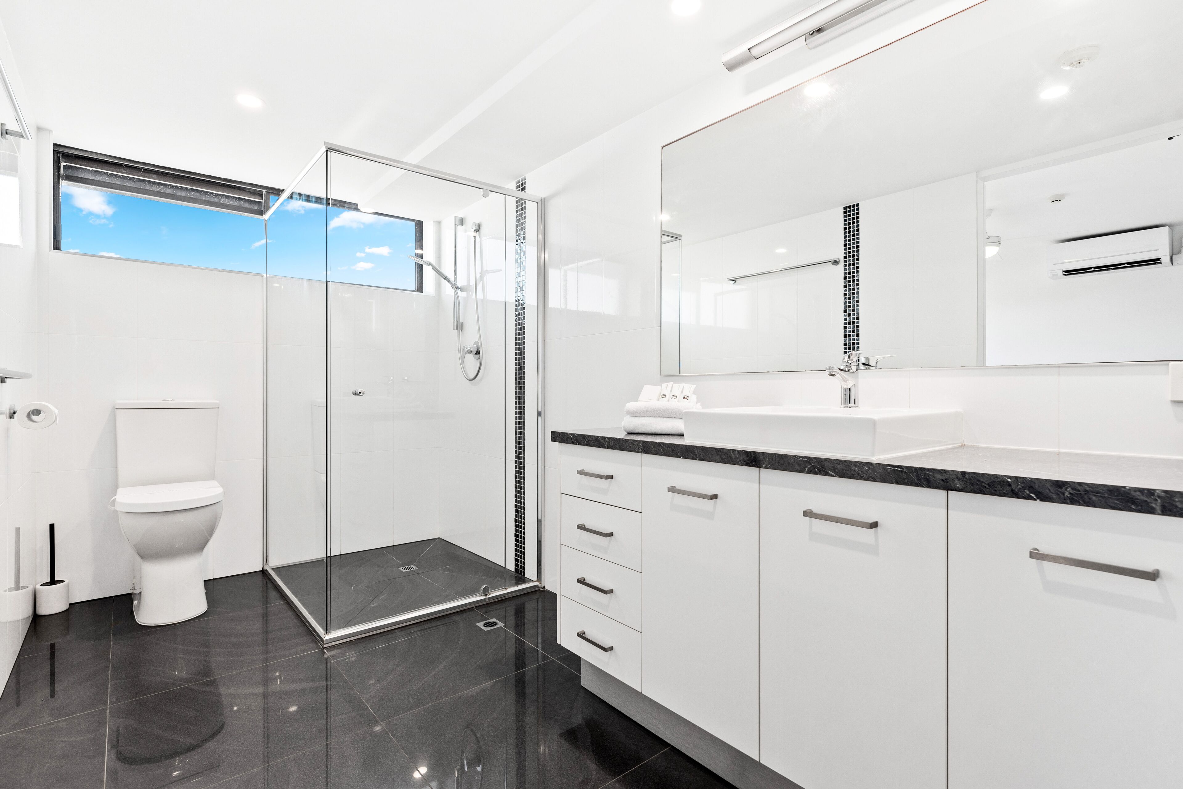 Un baño moderno con ducha envuelta en vidrio, inodoro blanco y un espejo grande sobre el lavamanos.