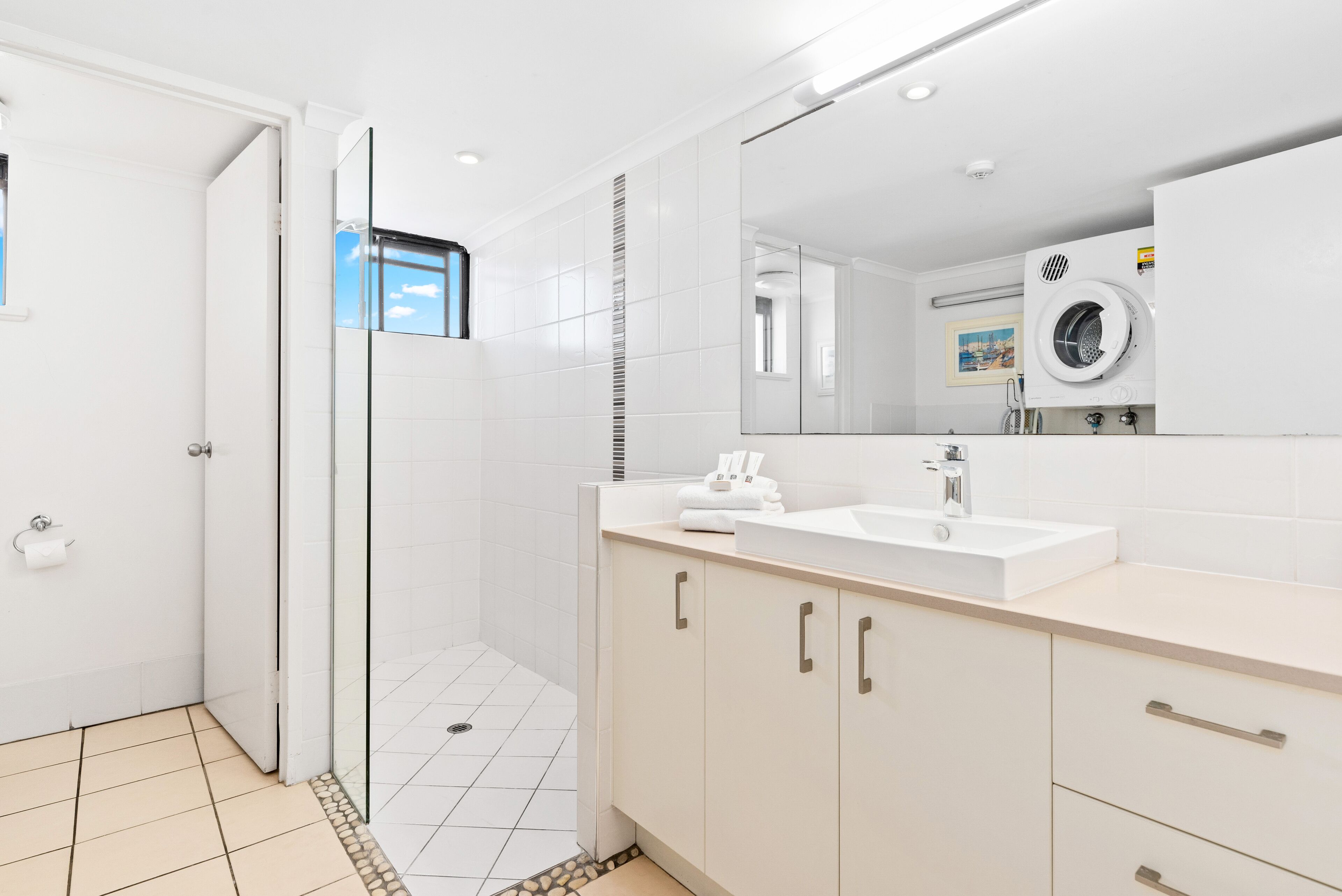A modern bathroom with a glass shower enclosure, a large mirror, a built-in washing machine, and a white sink.