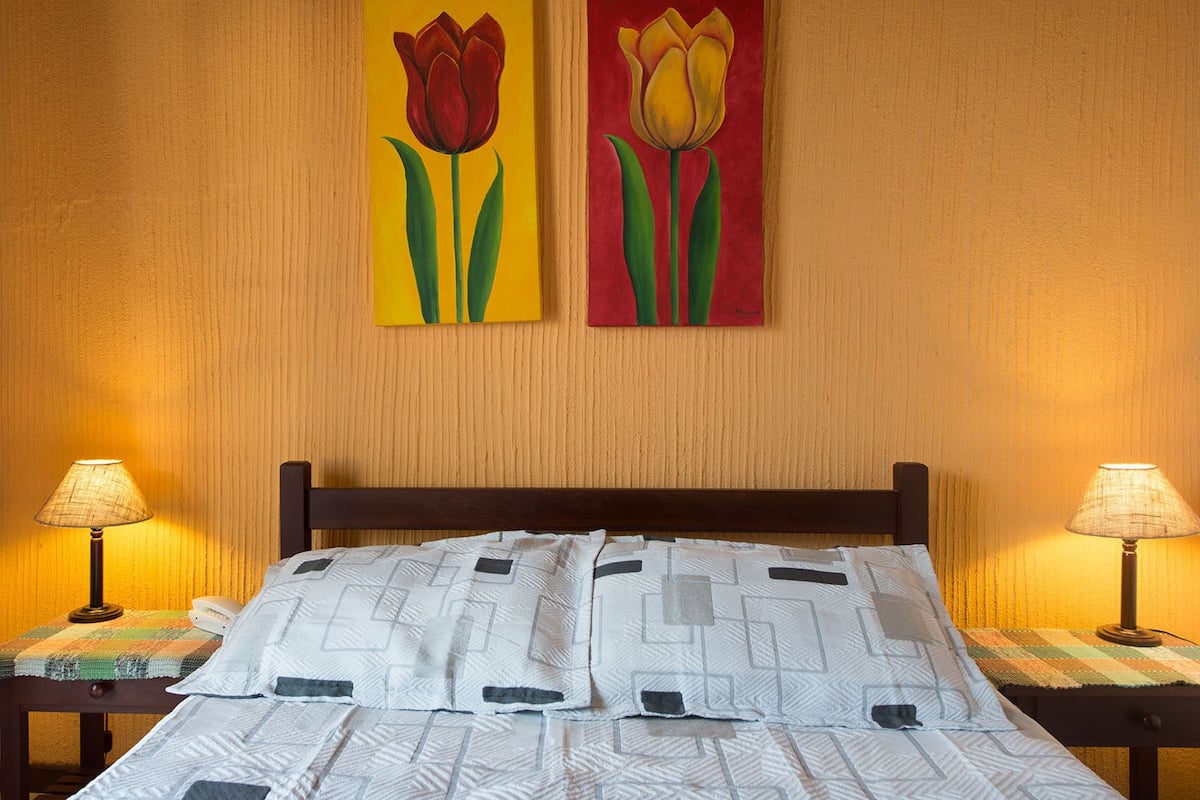 Quarto de hotel com cama, duas mesas de cabeceira, duas luminárias e duas pinturas de tulipas na parede.