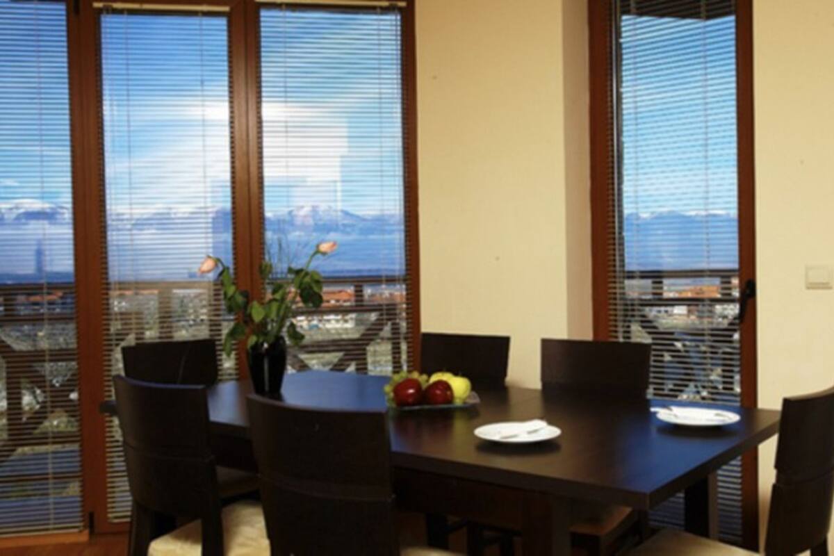Uma área de refeições com uma mesa posta para dois, cercada por cadeiras, e vista para a cidade através de amplas janelas.