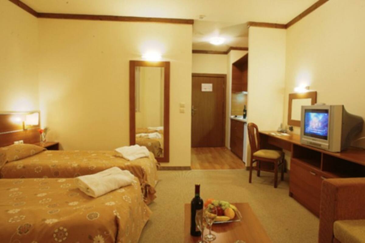 Quarto de hotel com duas camas, uma escrivaninha com televisão, uma cadeira e uma mesinha com uma garrafa e frutas.