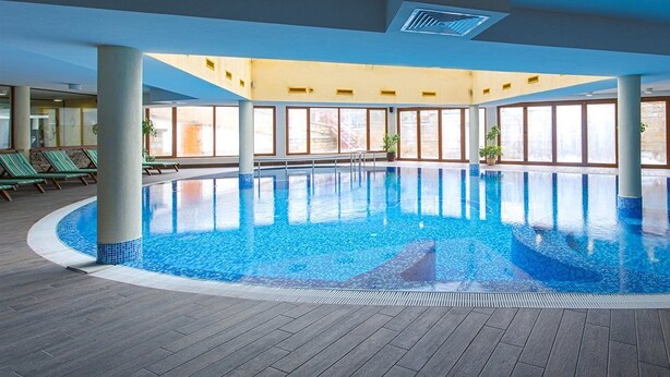 An indoor swimming pool with clear blue water, surrounded by large windows and a wooden deck. - Vihren Palace Ski & Spa Resort (Bansko)
