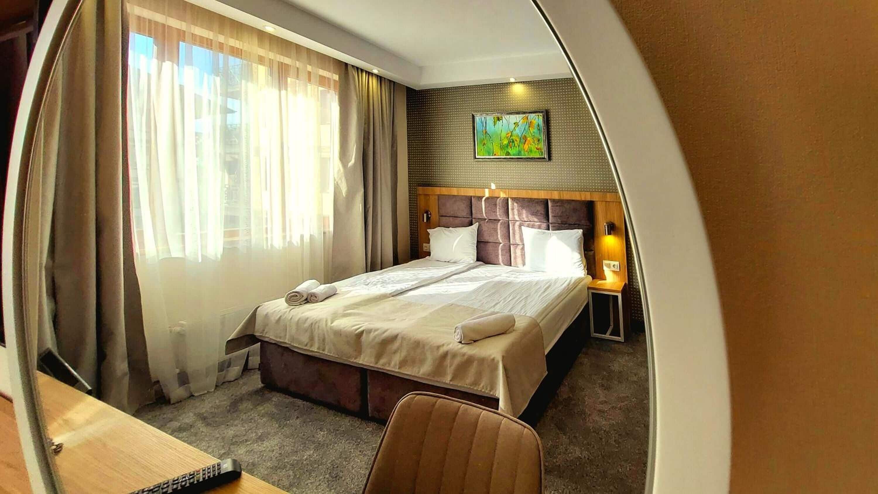 Une chambre d’hôtel avec un grand lit, une table de chevet avec une lampe, un tableau au mur et une vue sur l’extérieur à travers des rideaux transparents.