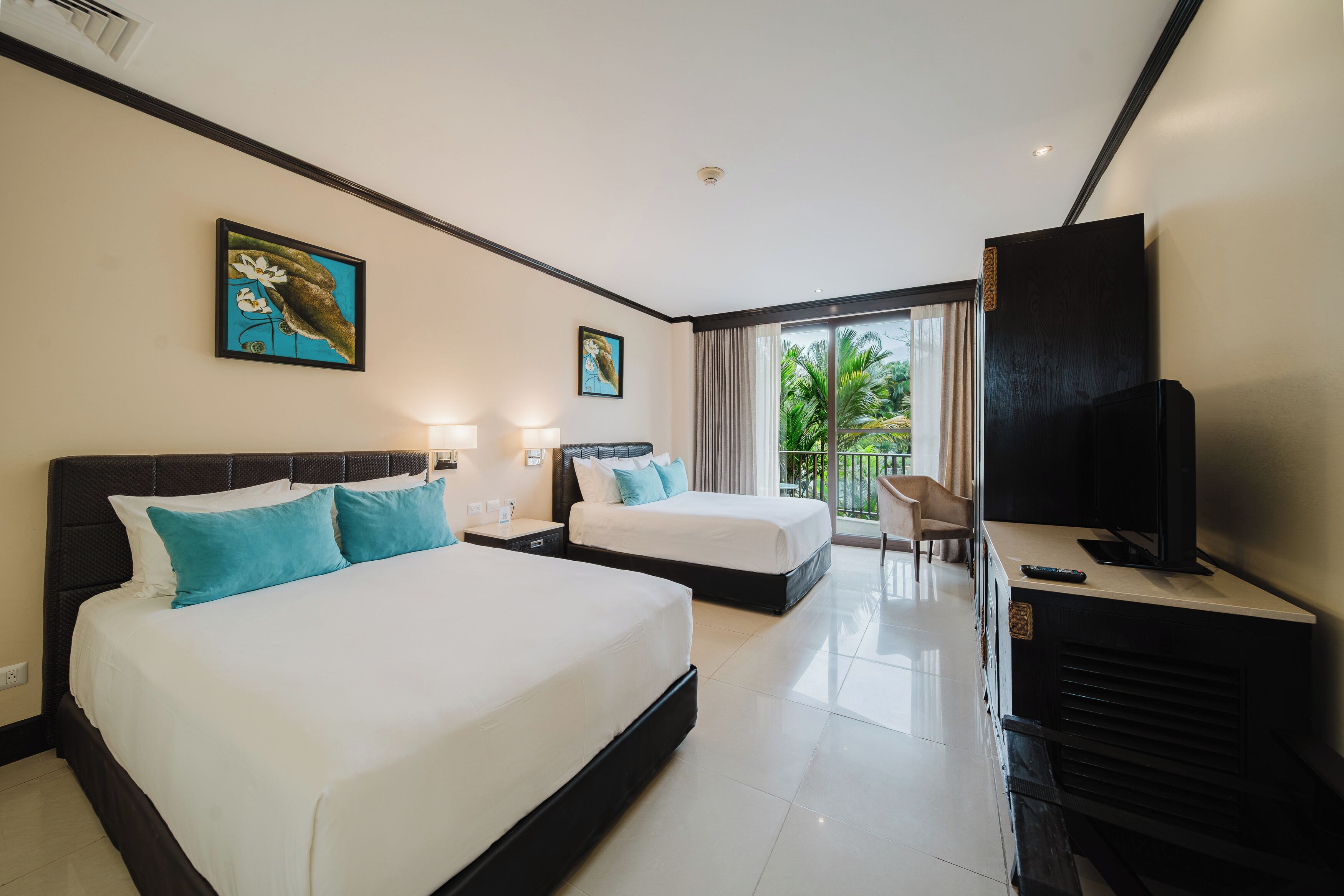 A hotel room with two beds, a TV, a desk, and a balcony with a view of greenery.