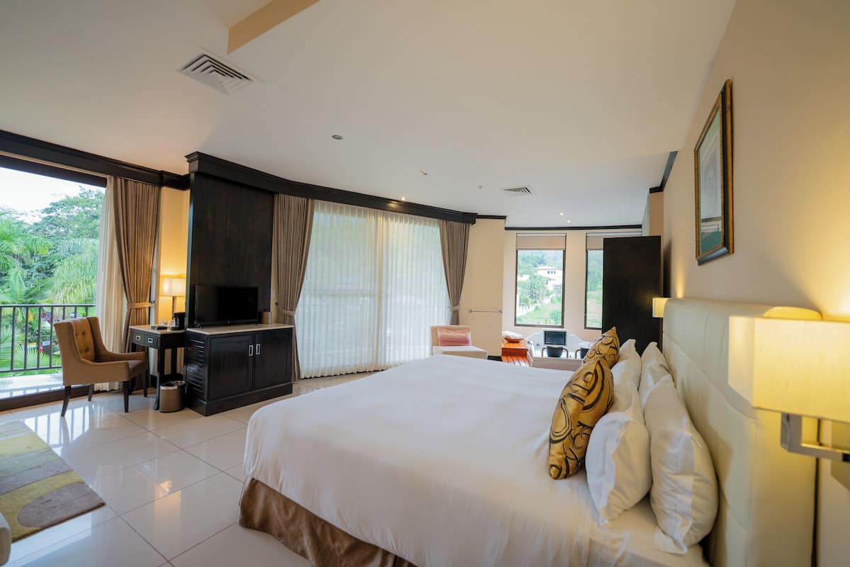 A hotel room with a large bed, a desk with a TV, a chair, and a balcony with curtains.