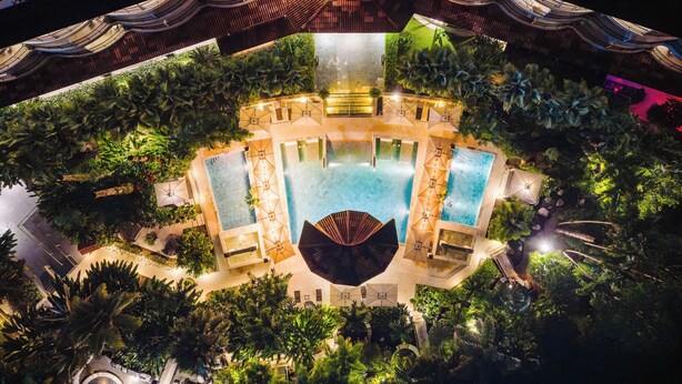 An aerial view of a resort with a central pool, surrounded by lush greenery and multiple levels of terraces.