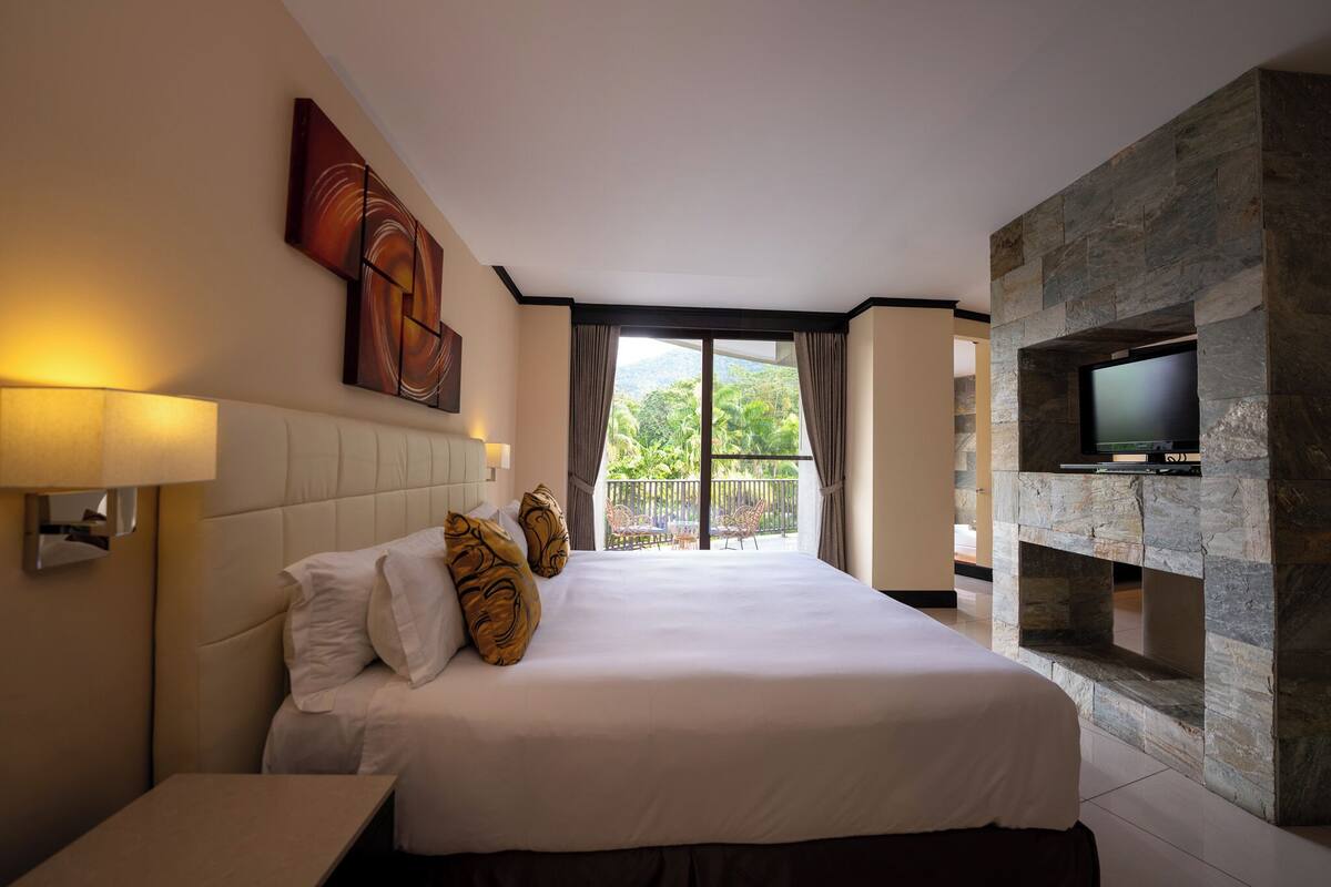 A hotel room with a large bed, a mounted TV, a stone fireplace, and a balcony with a view of greenery.