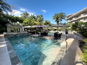 Zona de piscina con sillas de descanso y sombrillas, rodeada de palmeras y un edificio con balcones.