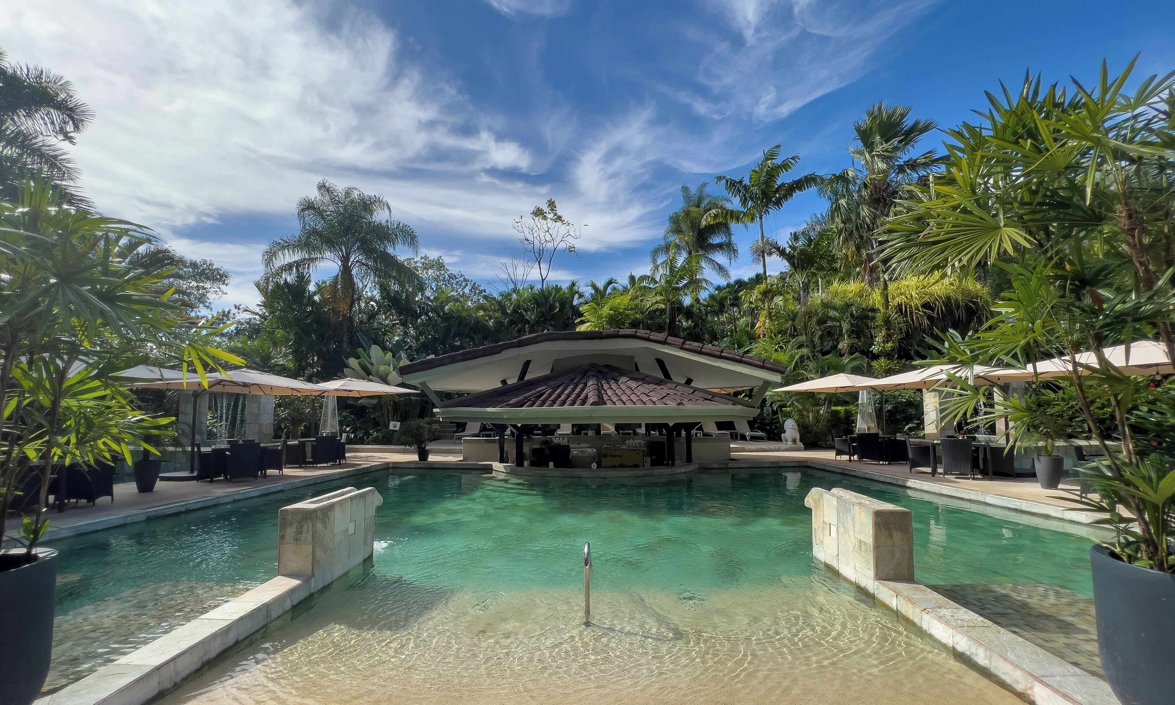 A clear pool with a central structure, surrounded by palm trees and outdoor seating.