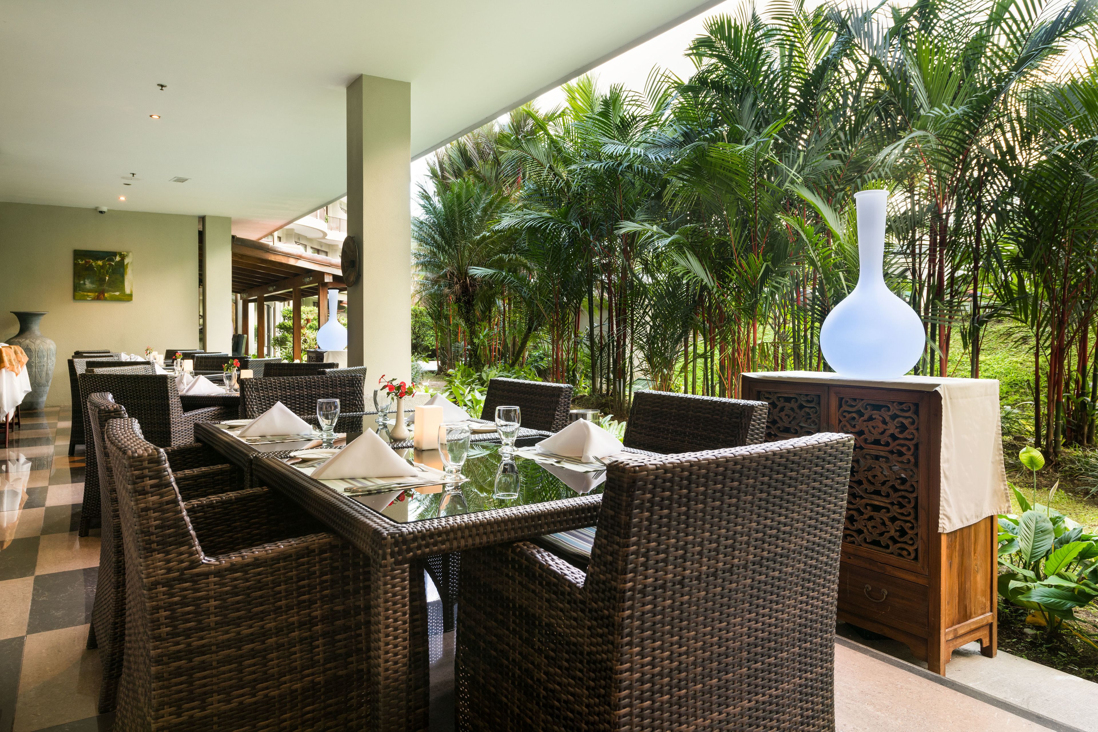 A restaurant with wicker furniture, a glass table, and a view of palm trees.