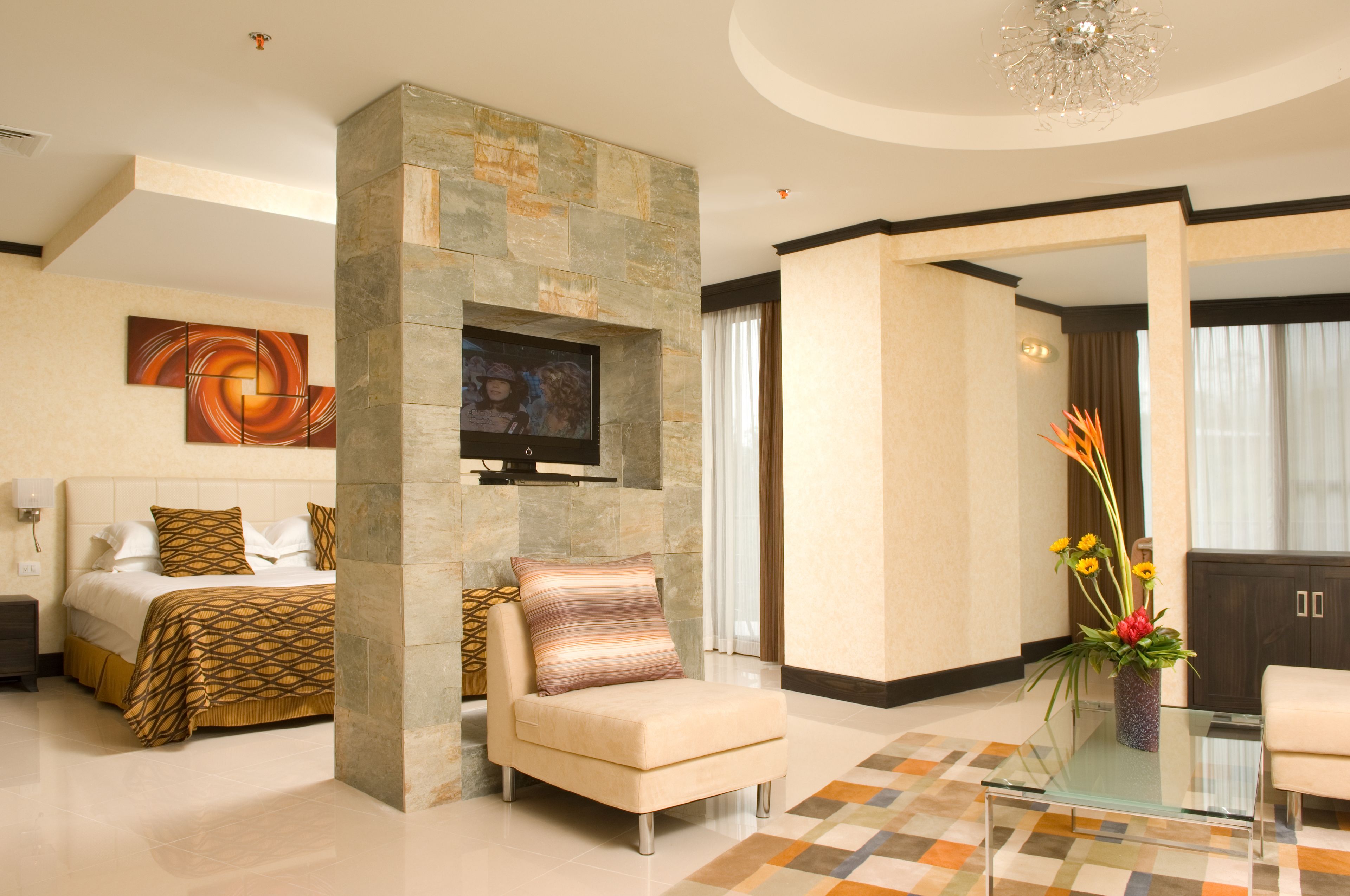 Quarto de hotel moderno com parede de detalhes em pedra, TV de tela plana, sofá confortável, mesa de centro de vidro e um vaso decorativo com flores.